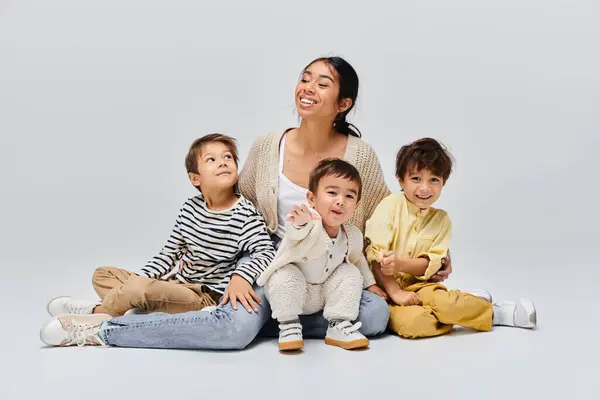 Una joven madre asiática se sienta en el suelo con sus hijos a su lado en un estudio sobre un fondo gris. - foto de stock