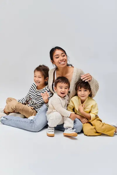 Una joven madre asiática se sienta tranquilamente en el suelo con sus hijos en un estudio sobre un fondo gris. - foto de stock