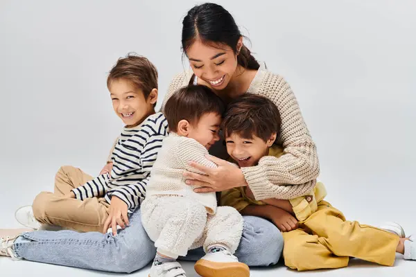 Una joven y pacífica madre asiática se sienta en el suelo, sosteniendo a sus hijos en un estudio con un fondo gris. - foto de stock