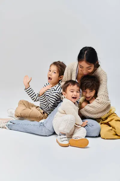 Una joven madre asiática sentada en el suelo rodeada de sus hijos en un estudio sobre un fondo gris. - foto de stock
