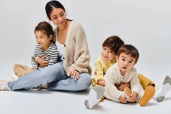 Joven madre asiática sentada en el suelo con sus hijos en un estudio sobre un fondo gris. - foto de stock
