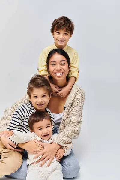 Una joven madre asiática posa cariñosamente con sus hijos en un estudio, sobre un fondo gris suave. - foto de stock