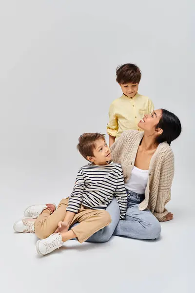 Una joven madre asiática está sentada en el suelo con sus hijos en un estudio sobre un fondo gris. - foto de stock