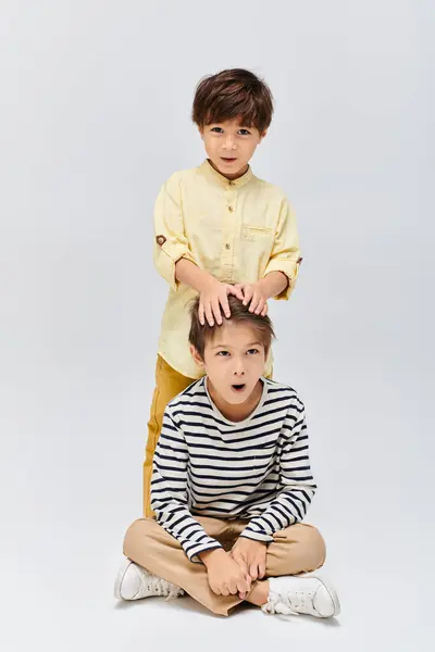 Two young boys seated on ground, hands atop their heads, lost in thought on a grey studio backdrop — Stock Photo