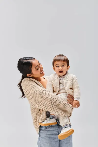Молода азіатська мати рве дитину на руках у студії на сірому фоні. — стокове фото