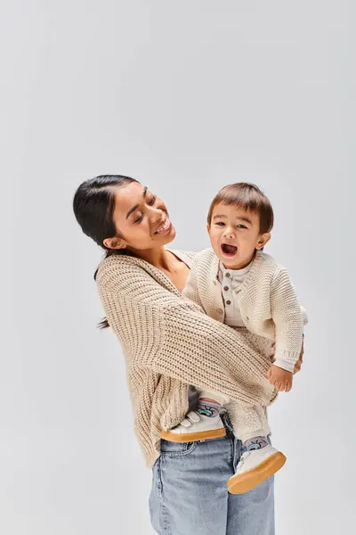A young Asian mother tenderly holds her baby in her arms, showing love and care in a studio setting against a grey background. — Stock Photo