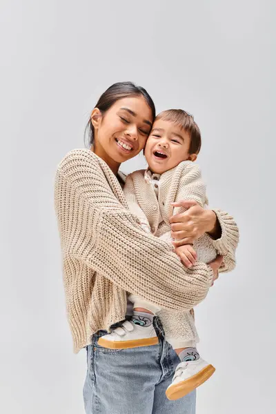 Una joven madre asiática acunando tiernamente a su bebé en sus brazos en un estudio sobre un fondo gris. - foto de stock