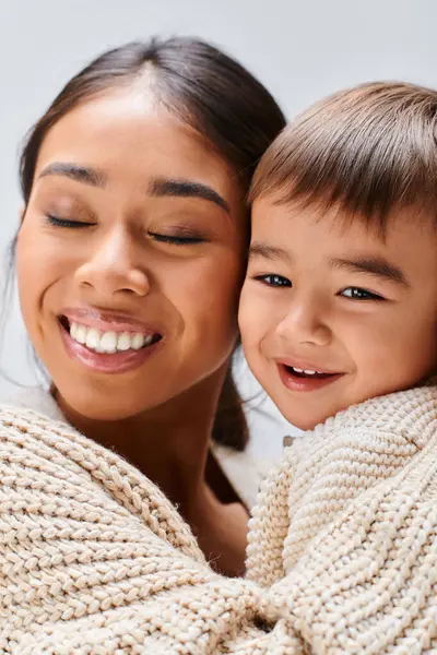 A young Asian mother lovingly holds her child in a studio setting against a grey background, close up — Stock Photo
