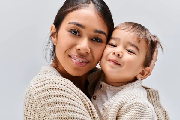 A young Asian mother tenderly holds her child in her arms in a studio setting against a grey background. — Stock Photo