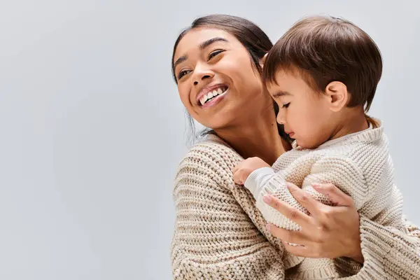 A young Asian mother cradles her child in a tender embrace against a grey backdrop. — Stock Photo