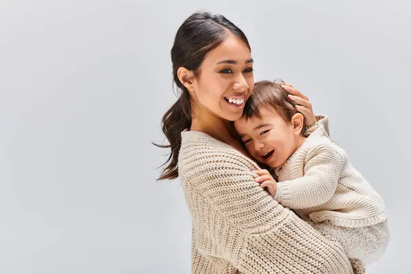 A young Asian mother tenderly embracing her child in a studio setting against a grey background. — Stock Photo