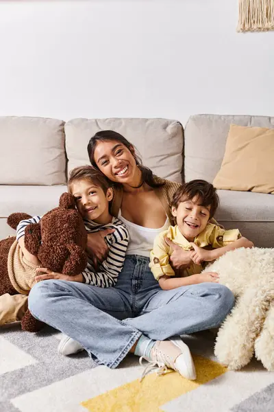 A young Asian mother sitting on the floor with her two children, accompanied by a teddy bear, in their cozy living room. — Stock Photo