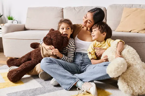 A young Asian mother sits on the floor with her two young sons and a teddy bear in a cozy living room setting. — Stock Photo