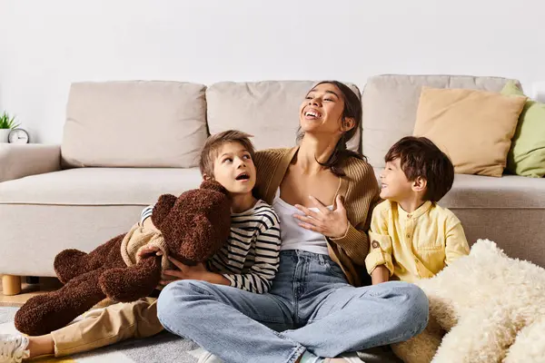A young Asian mother sitting on the floor with her two children and a teddy bear, creating a cozy family moment. — Stock Photo