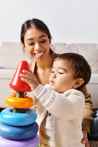 Una joven madre asiática juega alegremente con su pequeño hijo en el suelo de su acogedora sala de estar. - foto de stock