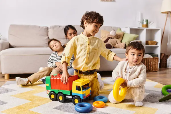 Un grupo de niños, incluidos los hijos pequeños de una joven madre asiática, están jugando felizmente con juguetes en una sala de estar cálida y acogedora. - foto de stock