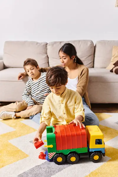 Groupe d'enfants, y compris la jeune mère asiatique, jouer avec un camion jouet dans un salon confortable rempli de rire. — Photo de stock