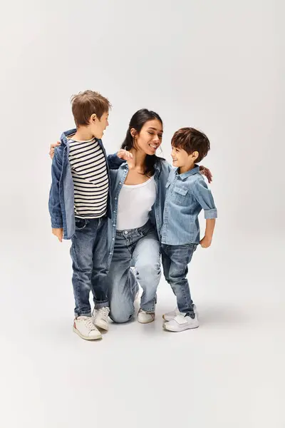 A young Asian mother and her two little sons standing together in a grey studio, all dressed in denim clothes. — Stock Photo