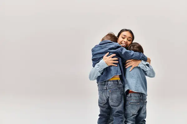 A young Asian mother and her little sons, wearing denim clothes, hugging each other in front of a white background. — Stock Photo