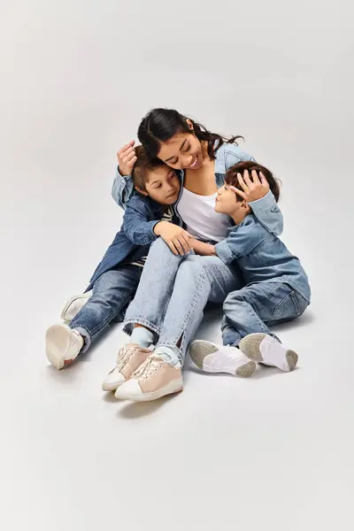 Una giovane madre asiatica e i suoi piccoli figli, tutti vestiti in denim, in posa insieme in uno studio grigio. — Foto stock
