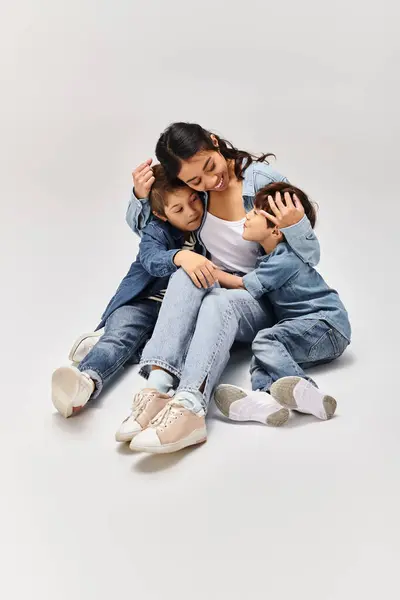 A young Asian mother and her little sons, all dressed in denim, posing together in a grey studio. — Stock Photo
