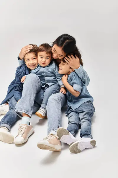 A young Asian mother and her little sons, all in denim clothes, creatively seated on top of each other in a grey studio setting. — Stock Photo