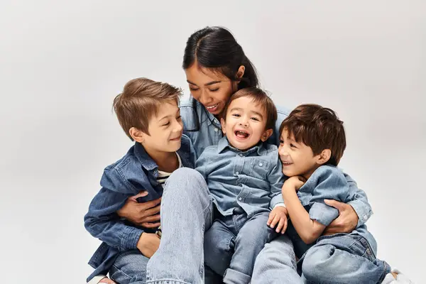 Un gruppo di bambini, tra cui una giovane madre asiatica e i suoi figli, vestiti di denim, si siedono uno sopra l'altro in uno studio grigio. — Foto stock