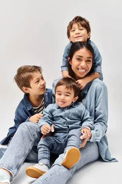 Un grupo de personas, una joven madre asiática y sus hijos pequeños, sentados uno encima del otro en un estudio gris, todos con ropa de mezclilla. - foto de stock