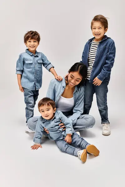 Una giovane madre asiatica e i suoi piccoli figli, tutti vestiti in denim, posano insieme in uno studio grigio. — Foto stock