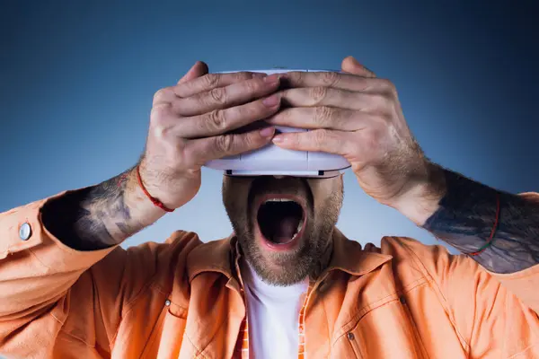 Un uomo con una camicia arancione tiene un oggetto bianco sopra la testa in uno studio mentre indossa un visore VR. — Foto stock