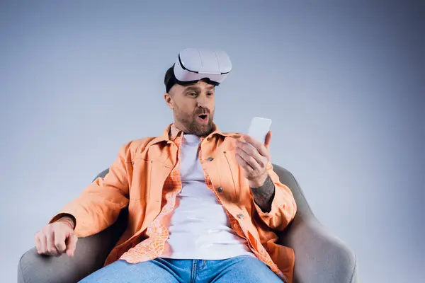 Un hombre inmerso en el mundo virtual, sentado en una silla con un teléfono celular en la mano, mezclando realidades en un entorno de estudio. - foto de stock