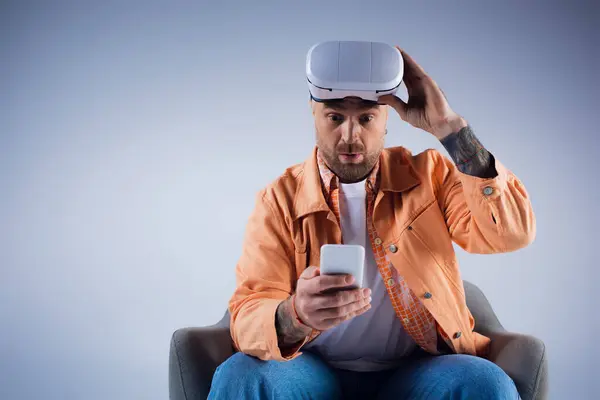 Um homem em uma cadeira segura um smartphone em um ambiente de estúdio, imerso no mundo do metaverso através de seu fone de ouvido VR. — Fotografia de Stock