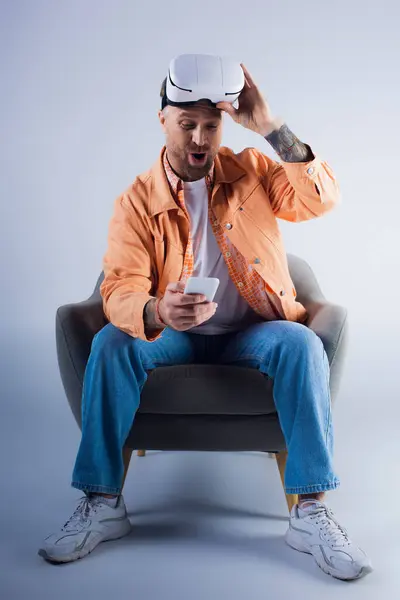 Un hombre se sienta en una silla mirando fijamente a un teléfono celular, profundamente en el pensamiento. - foto de stock