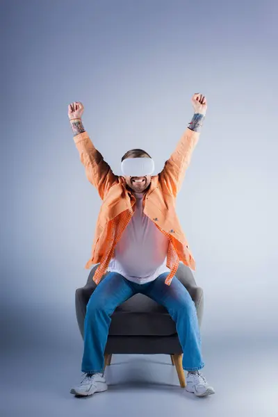 Чоловік у гарнітурі VR сидить на стільці з гербами, піднятими на святкування в студії. — стокове фото
