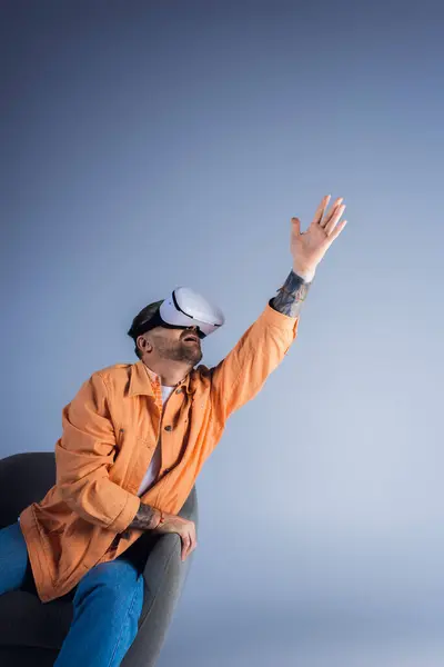 Un homme dans un casque VR est assis sur une chaise avec sa main levée, immergé dans un monde virtuel dans un cadre de studio. — Photo de stock