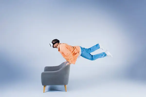 Um homem com uma camisa laranja e fones de ouvido vr mostra sua agilidade, equilibrando e realizando um truque em uma cadeira em um ambiente de estúdio. — Fotografia de Stock
