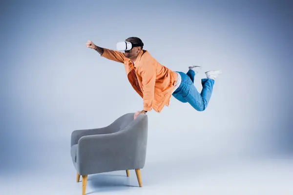 Un uomo con una camicia arancione in mostra un trucco che sfida la gravità mentre si bilancia su una sedia in un ambiente da studio. — Foto stock