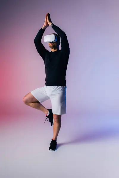 Un hombre con una camisa negra y pantalones cortos blancos practica una postura desafiante de yoga con equilibrio y control en un entorno de estudio. - foto de stock