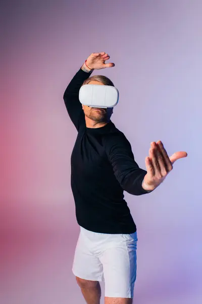 Un uomo in camicia nera e pantaloncini bianchi è sorprendente pose dinamiche in un ambiente di studio mentre immerso nel mondo della realtà virtuale. — Foto stock