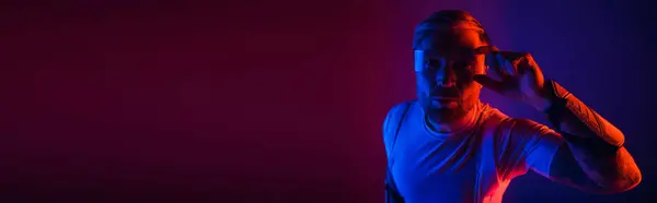 Um homem de óculos inteligentes fica confiante na frente de um vibrante fundo vermelho e azul em um estúdio. — Fotografia de Stock