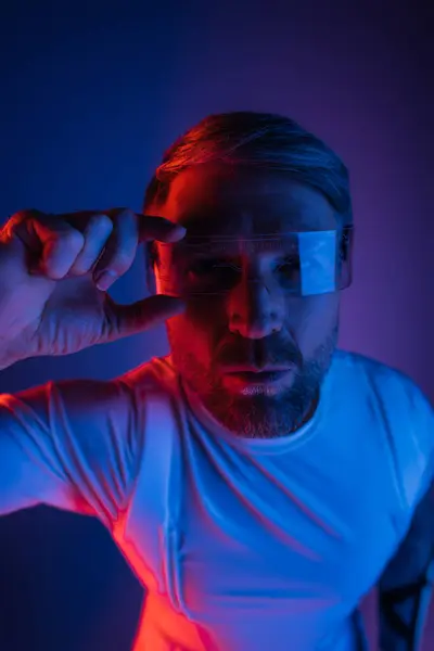 Un hombre con gafas inteligentes se para con confianza frente a un vibrante fondo rojo y azul en un entorno de estudio. - foto de stock