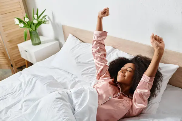Curly African American woman laying on bed with arms raised in pajamas, peaceful morning in bedroom. — Stock Photo