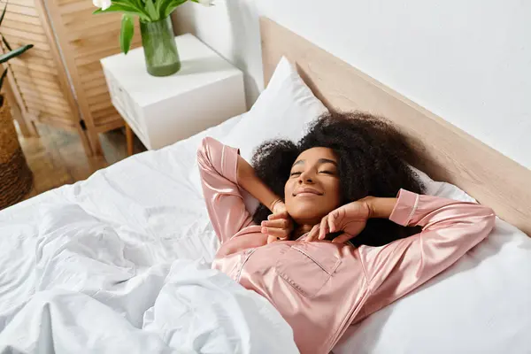 Eine lockige Afroamerikanerin im Pyjama, friedlich auf einem weißen Bett in einem sonnenbeschienenen Schlafzimmer liegend während des Morgens. — Stockfoto