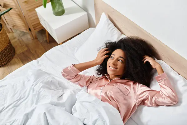 A curly African American woman in pajamas peacefully laying on top of a white bed in the morning. - foto de stock