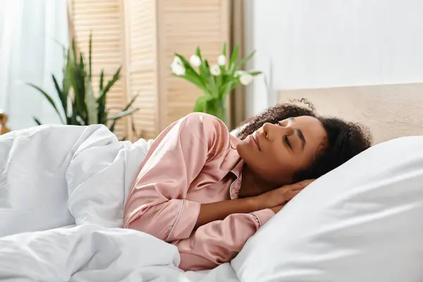 Eine lockige Afroamerikanerin im Schlafanzug entspannt sich morgens auf einem Bett mit weißen Laken. — Stockfoto