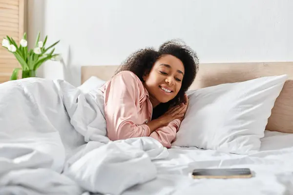 Riccio afroamericano donna in pigiama tranquillamente sdraiato su un letto con lenzuola bianche in un accogliente camera da letto. — Foto stock