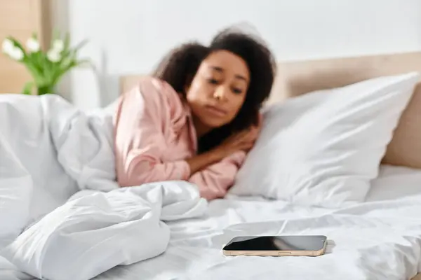 Une femme afro-américaine en pyjama se trouve au lit, absorbée dans son téléphone portable dans la douce lumière du matin. — Photo de stock