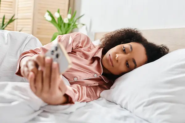 Lockige Afroamerikanerin im Schlafanzug liegt im Bett und hält morgens friedlich eine Fernbedienung in der Hand. — Stockfoto
