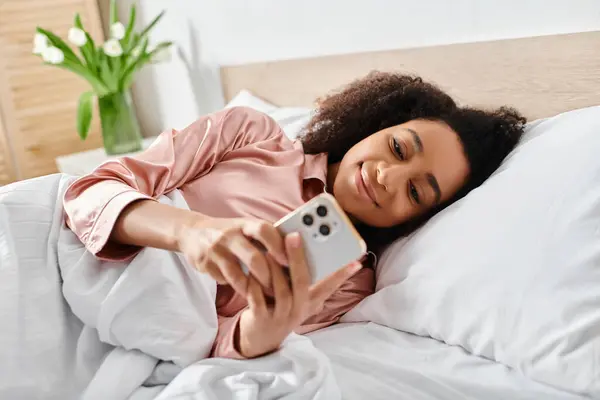 Curly African American woman in pajamas, laying in bed holding a cell phone in the morning. — Stock Photo