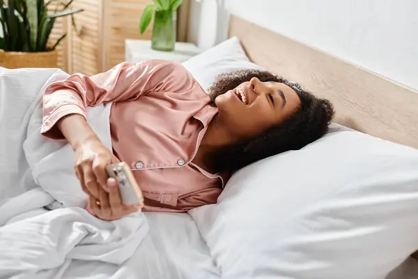 Femme afro-américaine bouclée en pyjama, allongée paisiblement sur le lit avec un téléphone portable à la main le matin. — Photo de stock